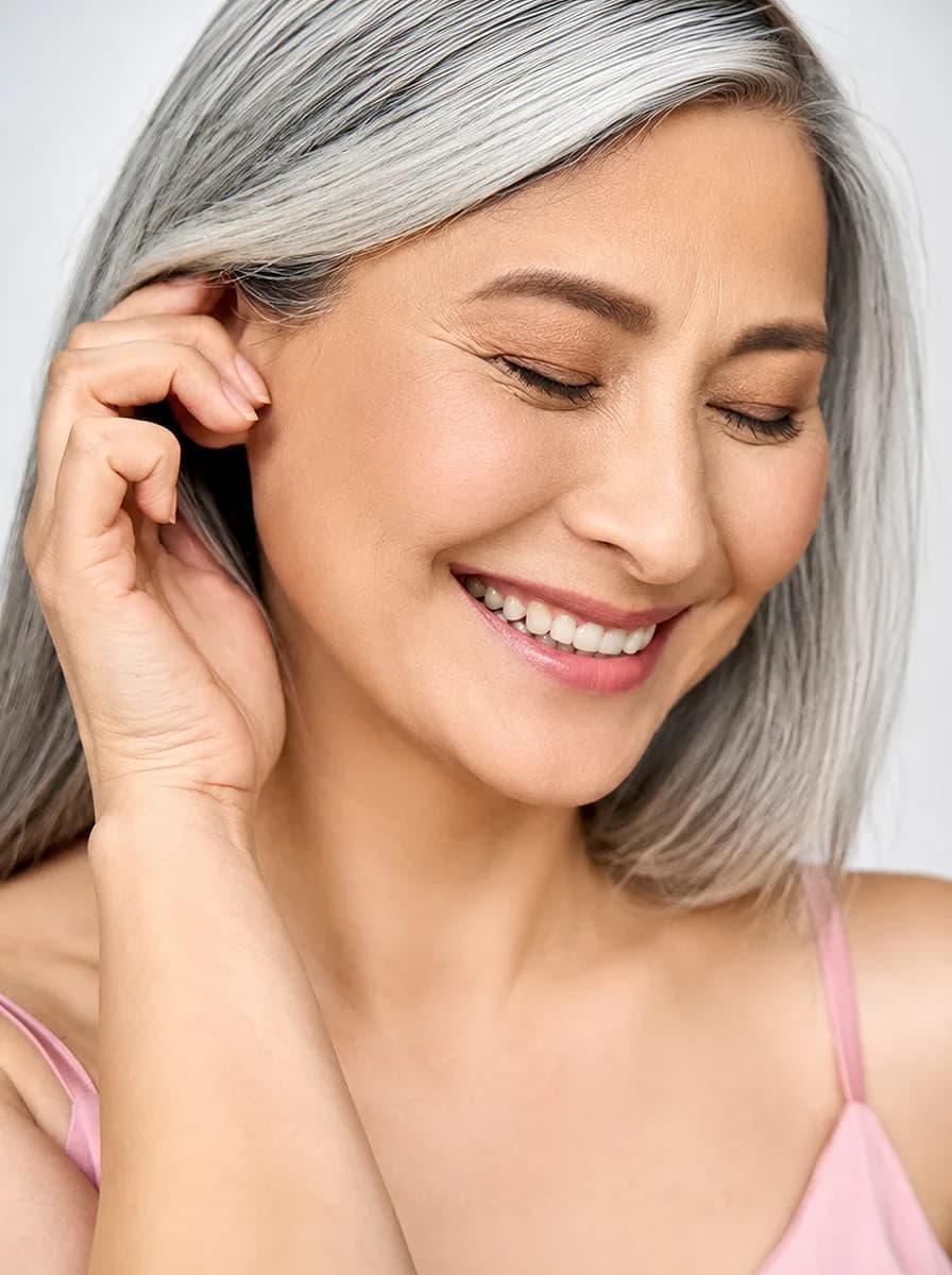 A silver-haired woman smiling with her eyes closed, tucking her hair behind her ear.