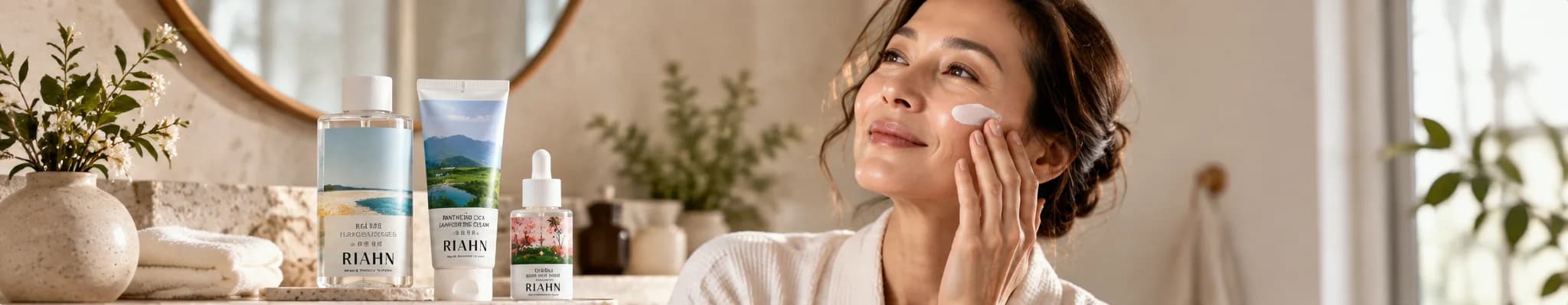 A woman in a white robe smoothing cream onto her cheek, three RIAHN pieces on the bathroom counter beside her.